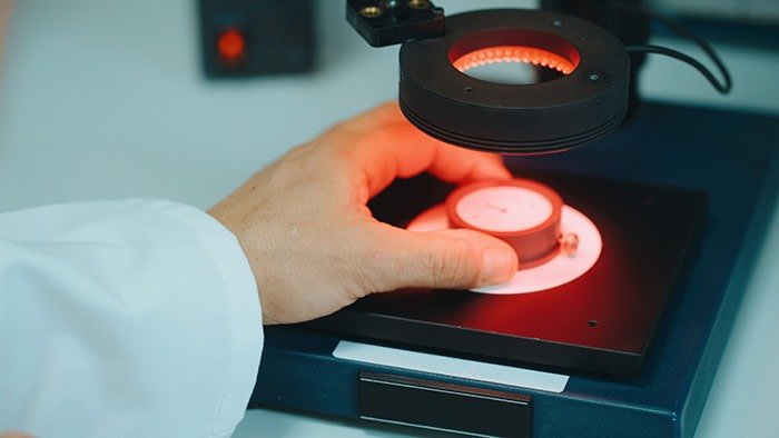 Hand holding a small watch component under a circular red inspection light.