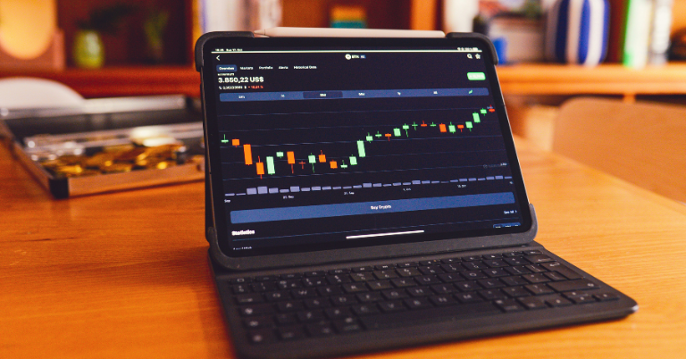 A tablet with a keyboard case displaying a candlestick chart for ETH cryptocurrency on a wooden table, with a stylus resting on top. The background shows a bookshelf with books, a vase, and a tray of gold coins.