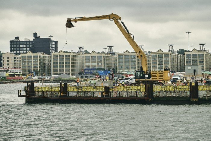 A large yellow crane sits on a pier with workers nearby, in front of industrial buildings along the Brooklyn waterfront.